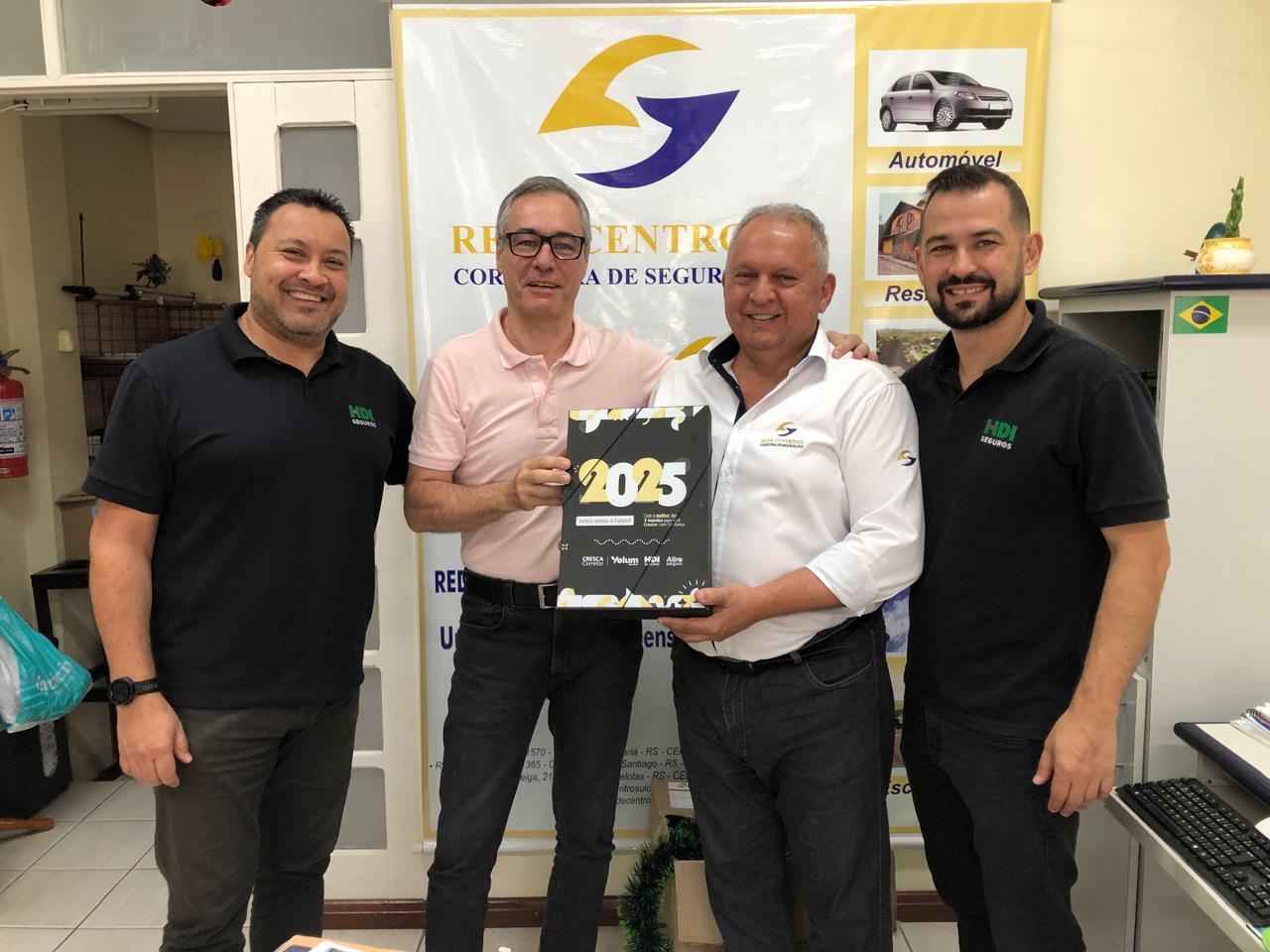 Na imagem aparecem quatro homens, sorridentes. Eles registram a renovação da parceria para 2025. Da esquerda para direita, está Gerente Regional Sucursal da HDI Seguros - Santa Cruz do Sul, Edmilson Cesar Alves, ele tem os cabelos pretos, pele clara e veste uma camiseta preta com a logo da HDI e uma calça preta, ao lado dele ao centro, estão os sócios-fundadores da Rede Centrosul, Glevander Ruviaro da Silva e Roberval Bresolin. Ruviaro, tem cabelos castanho claro e está de óculos com armação escura. Ele veste uma camiseta de cor rosa claro e calças pretas e segura com a mão direita, uma placa que está escrito 2025 com números grandes. Ao lado dele, segurando a placa com a mão esquerda, está Bresolin, ele tem cabelos grisalhos e veste camisa branca, de magas longas, com a logomarca da rede e calças pretas. Ao lado dele, no canto direito da imagem, está o gerente de Contas de Santa Maria da HDI Seguros, Arion Silveira Corrêa, ele tem cabelos pretos, barba e veste a camiseta preta com a logo da HDI e calças pretas. Eles estão em frente ao banner da Rede Centrosul Corretora de Seguros.